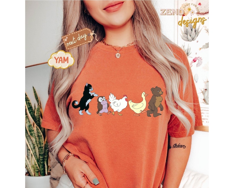 May include: Burnt orange t-shirt featuring a line of cartoon animals: a black cat, an owl, a white chicken, a yellow duck, and a brown bear. The shirt has the word "YAM" on a cloud-shaped tag and the words "next day" on a brown tag.
