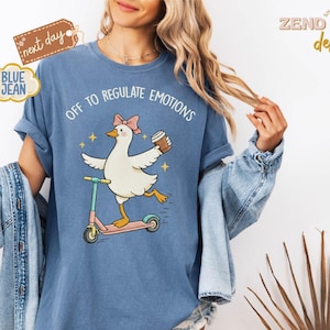 Puede incluir: Una camiseta azul con el gráfico de una oca blanca con un lazo rosa, montada en un patinete y sosteniendo un vaso de café. El texto sobre la oca dice "OFF TO REGULATE EMOTIONS". La camiseta se muestra con una chaqueta vaquera encima.