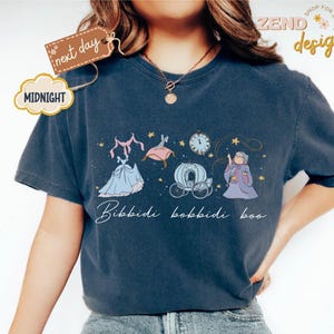 May include: Navy blue t-shirt featuring a Cinderella-themed graphic. The design includes a blue gown, a carriage, a clock, and the text "Bibbidi Bobbidi Boo." The shirt has a relaxed fit and short sleeves.