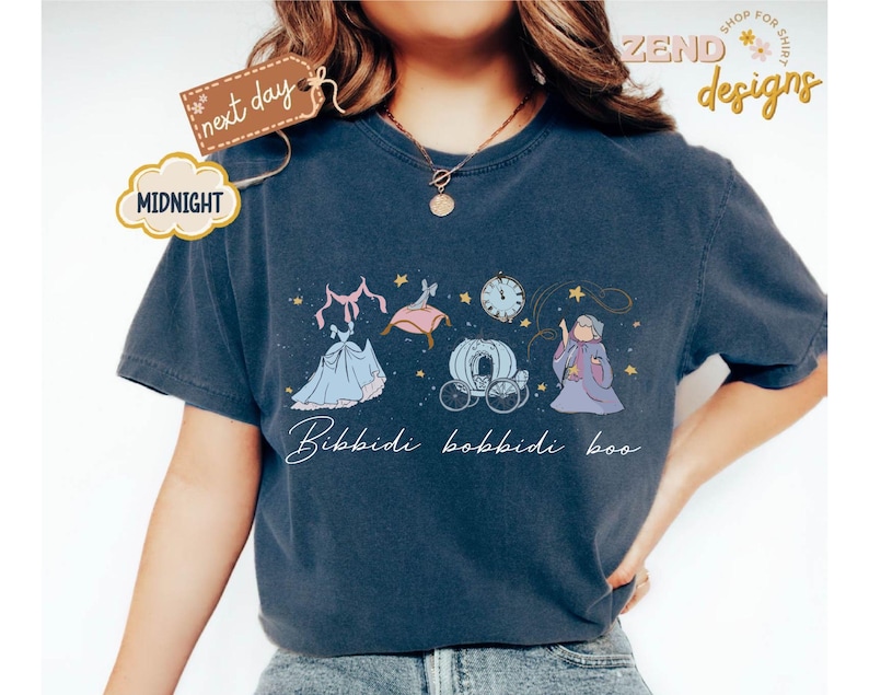 May include: Navy blue t-shirt featuring a Cinderella-themed graphic. The design includes a blue gown, a carriage, a clock, and the text "Bibbidi Bobbidi Boo." The shirt has a relaxed fit and short sleeves.
