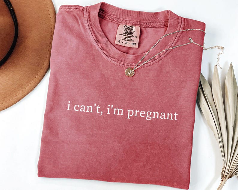May include: A red-orange Comfort Colors t-shirt with the text "i can't, i'm pregnant" in white. A gold necklace with a small pendant and a brown hat are also in the image.