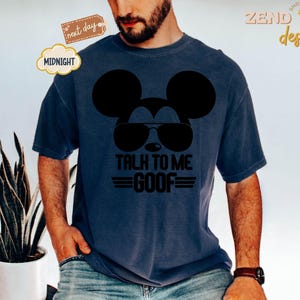 Puede incluir: Una camiseta de manga corta azul oscuro con un gr&aacute;fico negro de la silueta de un rat&oacute;n con gafas de aviador. Debajo del gr&aacute;fico, el texto dice TALK TO ME GOOF en letras may&uacute;sculas negras en negrita con l&iacute;neas horizontales a los lados.