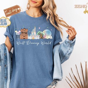 May include: Blue jean-colored t-shirt featuring a colorful illustration of Walt Disney World landmarks. The design includes Cinderella's castle, Spaceship Earth, and other iconic structures. The words "Walt Disney World" are written in a script font below the illustration.