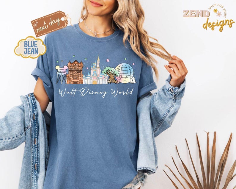 May include: Blue jean-colored t-shirt featuring a colorful illustration of Walt Disney World landmarks. The design includes Cinderella's castle, Spaceship Earth, and other iconic structures. The words "Walt Disney World" are written in a script font below the illustration.