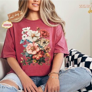 Puede incluir: Camiseta de color carmes&iacute; con un dise&ntilde;o de ramo floral. El ramo incluye flores blancas, rosas y melocot&oacute;n con hojas verdes. La camiseta tiene mangas cortas y un corte holgado. El texto "next day" y "Crimson" son visibles.