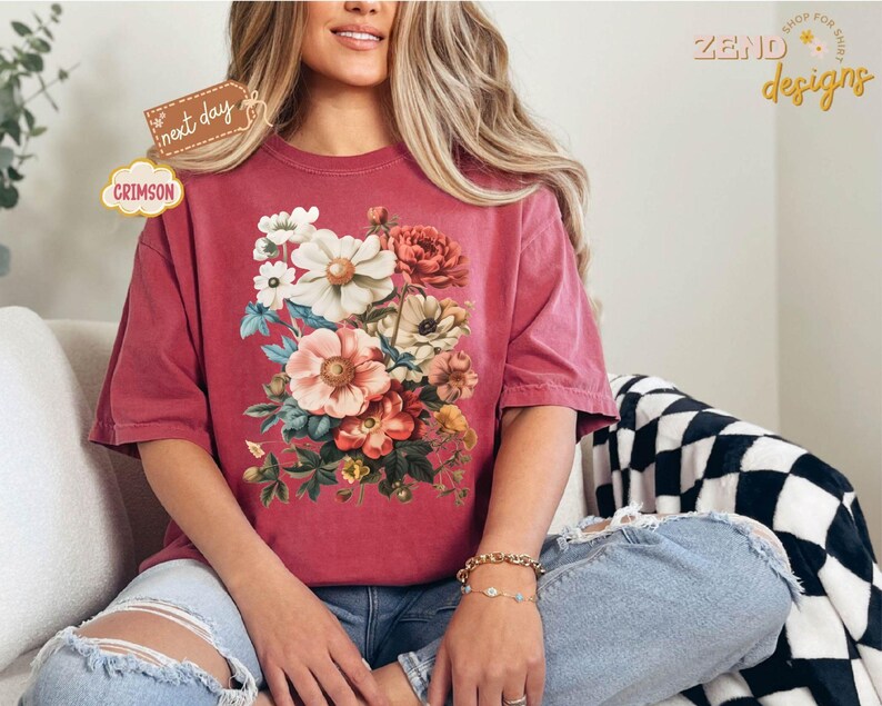 Puede incluir: Camiseta de color carmes&iacute; con un dise&ntilde;o de ramo floral. El ramo incluye flores blancas, rosas y melocot&oacute;n con hojas verdes. La camiseta tiene mangas cortas y un corte holgado. El texto "next day" y "Crimson" son visibles.