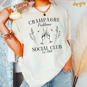 May include: A white t-shirt with a black graphic design that says "Champagne Problems Social Club Est. 1989". The graphic features two champagne glasses clinking together with a floral design.
