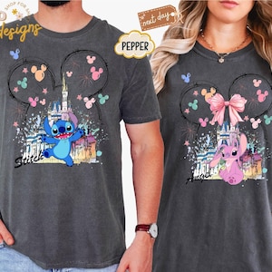 May include: Two charcoal gray t-shirts featuring Disney's Stitch and Angel characters. Each shirt has a castle graphic within a Mickey Mouse ear outline, with the character's name below. The Stitch shirt has blue accents, while the Angel shirt has pink.