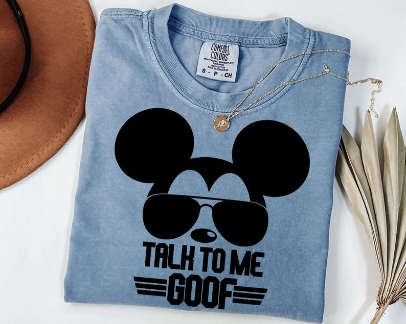 Puede incluir: Una camiseta azul claro de cuello redondo con un gr&aacute;fico negro de la silueta de un rat&oacute;n con gafas de sol. El texto debajo del gr&aacute;fico dice TALK TO ME GOOF con l&iacute;neas horizontales a los lados. Incluye la etiqueta de marca Comfort Colors.