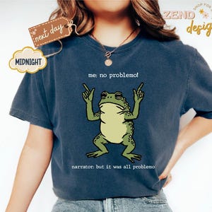Puede incluir: Camiseta azul marino con una ilustraci&oacute;n de rana verde y el texto "me: no problemo!" y "narrator: but it was all problemo". La camiseta tiene un corte relajado, ideal para uso diario.