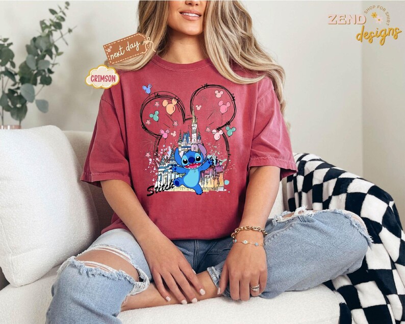 May include: Crimson-colored t-shirt featuring a graphic of the character Stitch from Lilo & Stitch, with a castle and Mickey Mouse ears design. The shirt has a relaxed fit and is worn with blue jeans. The word "Sweet" is printed on the shirt.