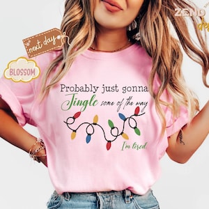 Pode incluir: Camiseta rosa claro com a frase "Probably just gonna Jingle some of the way. I'm tired." Uma sequ&ecirc;ncia de luzes de Natal est&aacute; abaixo do texto. A camiseta tem mangas curtas.