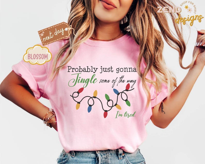 Pode incluir: Camiseta rosa claro com a frase "Probably just gonna Jingle some of the way. I'm tired." Uma sequ&ecirc;ncia de luzes de Natal est&aacute; abaixo do texto. A camiseta tem mangas curtas.