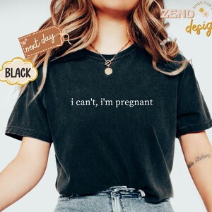 May include: Black t-shirt with the phrase "i can't, i'm pregnant" in white lettering. The shirt has short sleeves and a crew neck. A tag that says "next day" is attached to the shirt.