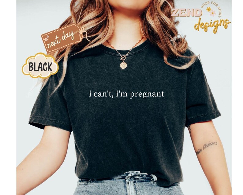 May include: Black t-shirt with the phrase "i can't, i'm pregnant" in white lettering. The shirt has short sleeves and a crew neck. A tag that says "next day" is attached to the shirt.