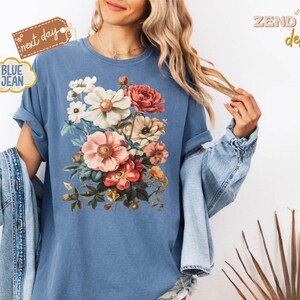 Puede incluir: Camiseta azul vaquera con un dise&ntilde;o de ramo de flores en tonos rosas, rojos, blancos y amarillos. La camiseta tiene las mangas enrolladas y se ven las palabras "next day" y "blue jean".