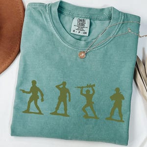 Puede incluir: Una camiseta verde agua Comfort Colors con un gr&aacute;fico de cuatro soldados de juguete verdes en varias poses. La camiseta tiene cuello redondo y mangas cortas. Un collar dorado con un colgante est&aacute; sobre la camiseta.