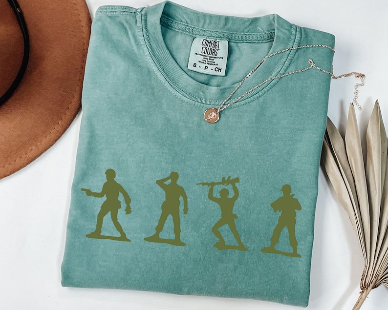 Puede incluir: Una camiseta verde agua Comfort Colors con un gr&aacute;fico de cuatro soldados de juguete verdes en varias poses. La camiseta tiene cuello redondo y mangas cortas. Un collar dorado con un colgante est&aacute; sobre la camiseta.