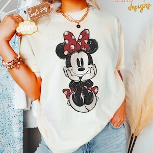 May include: A white t-shirt with a vintage-style illustration of Minnie Mouse. Minnie is wearing a red bow with white polka dots and a red skirt with white polka dots. She is smiling and has her hands on her chin.