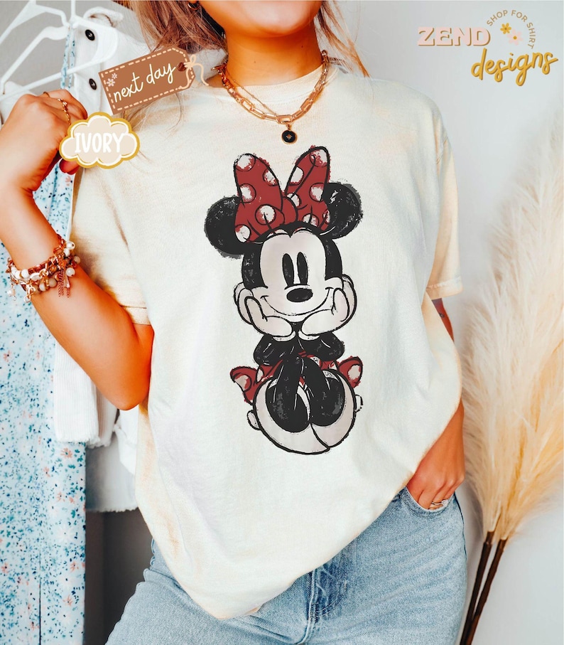 May include: A white t-shirt with a vintage-style illustration of Minnie Mouse. Minnie is wearing a red bow with white polka dots and a red skirt with white polka dots. She is smiling and has her hands on her chin.
