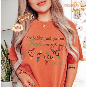 Pode incluir: Camiseta laranja com o texto "Probably just gonna Jingle some of the way, I'm tired" e uma sequ&ecirc;ncia de luzes de Natal coloridas. A camiseta tem um caimento folgado e &eacute; feita de um material macio.