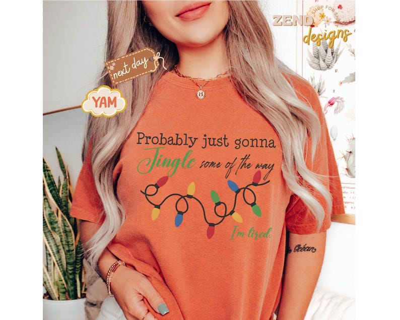 Pode incluir: Camiseta laranja com o texto "Probably just gonna Jingle some of the way, I'm tired" e uma sequ&ecirc;ncia de luzes de Natal coloridas. A camiseta tem um caimento folgado e &eacute; feita de um material macio.