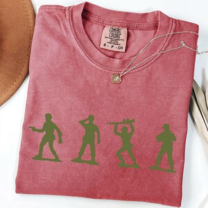 Puede incluir: Una camiseta roja Comfort Colors con cuatro siluetas de soldados de juguete verdes. La camiseta tiene cuello redondo y una peque&ntilde;a etiqueta en la parte superior derecha. Un collar plateado con un colgante est&aacute; sobre la camiseta.