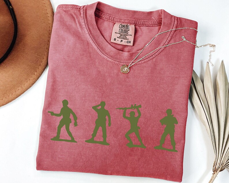 Puede incluir: Una camiseta roja Comfort Colors con cuatro siluetas de soldados de juguete verdes. La camiseta tiene cuello redondo y una peque&ntilde;a etiqueta en la parte superior derecha. Un collar plateado con un colgante est&aacute; sobre la camiseta.