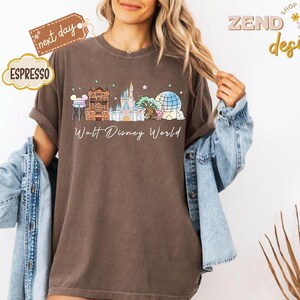 May include: Espresso-colored t-shirt featuring a Walt Disney World design. The graphic includes iconic park landmarks in a colorful, whimsical style. The shirt has rolled-up sleeves and a relaxed fit.