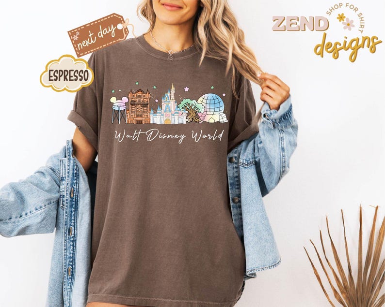 May include: Espresso-colored t-shirt featuring a Walt Disney World design. The graphic includes iconic park landmarks in a colorful, whimsical style. The shirt has rolled-up sleeves and a relaxed fit.