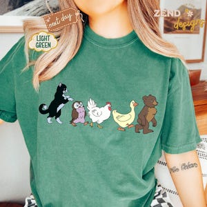 May include: A light green t-shirt featuring a cartoon illustration of a black cat, owl, chicken, duck, and bear walking together. The shirt has the text "ZEND designs" and "next day" and "light green" on the shirt.