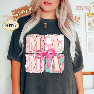 Puede incluir: Una camiseta negra con un dise&ntilde;o gr&aacute;fico de la palabra "MAMA" en letras rosas y blancas rodeada de dise&ntilde;os florales y de lazos.