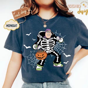 K&ouml;nnte beinhalten: Dunkelblaues T-Shirt mit einem Halloween-Motiv von Buzz Lightyear. Die Grafik zeigt Buzz Lightyear in einem Skelettkost&uuml;m, der einen K&uuml;rbis mit sich tr&auml;gt. Wei&szlig;e Spinnweben und Flederm&auml;use sind ebenfalls im Design enthalten. Das Shirt hat den Text "Midnight" und "next day".