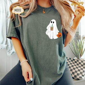 Pu&ograve; includere: Una maglietta verde scuro con un fantasma bianco che tiene una tazza di caff&egrave; e una piccola zucca arancione. Il fantasma ha un viso amichevole e indossa un lenzuolo bianco.
