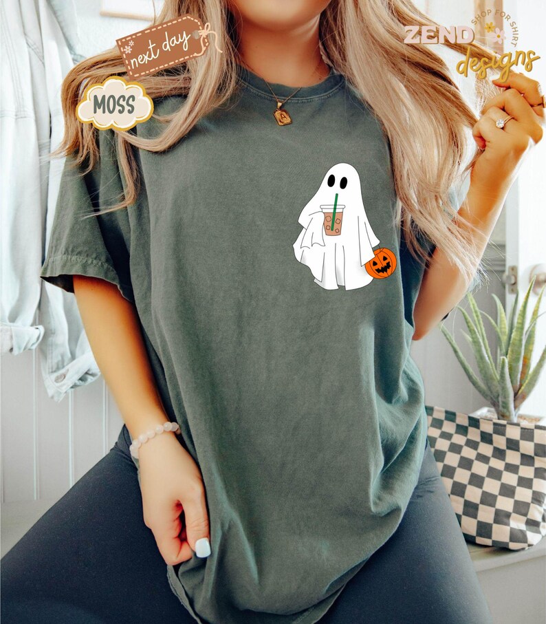 Pu&ograve; includere: Una maglietta verde scuro con un fantasma bianco che tiene una tazza di caff&egrave; e una piccola zucca arancione. Il fantasma ha un viso amichevole e indossa un lenzuolo bianco.
