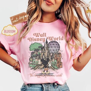 以下が含まれることがあります： ウォルト・ディズニー・ワールドのイラストが描かれたピンク色のTシャツ。ミッキーマウスと象徴的なランドマークが特徴です。「ウォルト・ディズニー・ワールド」と「The happiest place on earth」の文字が含まれています。