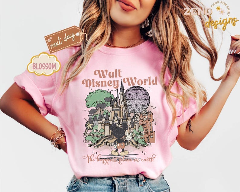 以下が含まれることがあります： ウォルト・ディズニー・ワールドのイラストが描かれたピンク色のTシャツ。ミッキーマウスと象徴的なランドマークが特徴です。「ウォルト・ディズニー・ワールド」と「The happiest place on earth」の文字が含まれています。