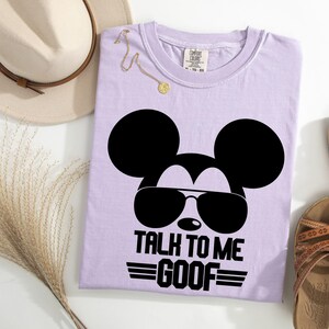 Puede incluir: Una camiseta de color morado claro con un gr&aacute;fico negro de una cabeza de rat&oacute;n con gafas de sol y el texto "TALK TO ME GOOF" impreso debajo. La camiseta tiene cuello redondo y una etiqueta de la marca Comfort Colors en el cuello.