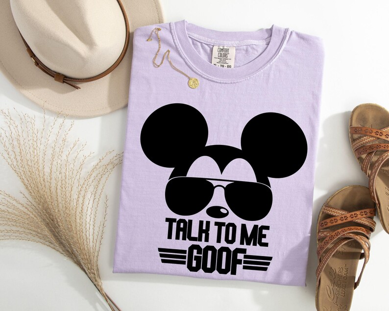 Puede incluir: Una camiseta de color morado claro con un gr&aacute;fico negro de una cabeza de rat&oacute;n con gafas de sol y el texto "TALK TO ME GOOF" impreso debajo. La camiseta tiene cuello redondo y una etiqueta de la marca Comfort Colors en el cuello.