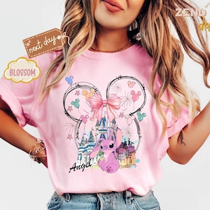 May include: Pink t-shirt featuring a whimsical design with a castle, fireworks, and a cartoon character. The design includes a large bow, Mickey Mouse ears, and the word "Angel". The shirt has short sleeves.