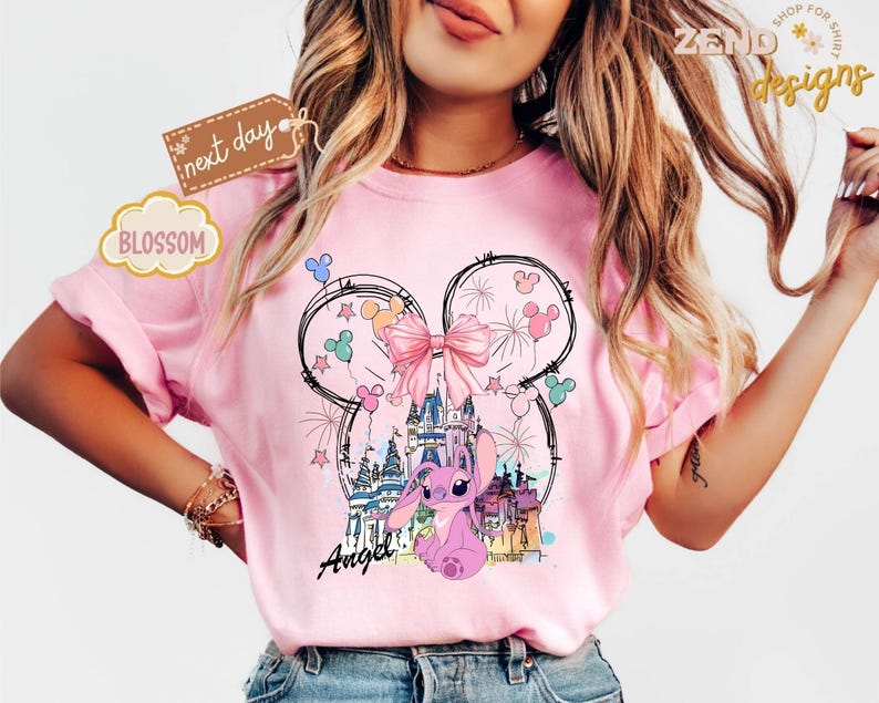 May include: Pink t-shirt featuring a whimsical design with a castle, fireworks, and a cartoon character. The design includes a large bow, Mickey Mouse ears, and the word "Angel". The shirt has short sleeves.