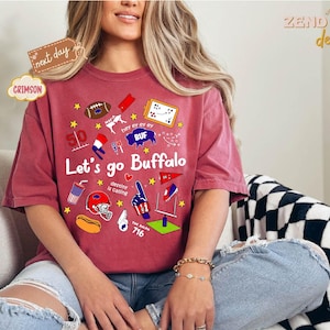 May include: Crimson-colored t-shirt with the text "Let's go Buffalo" and various graphics, including a football, stars, and a helmet. The shirt also features the text "destiny is calling" and the number "716".