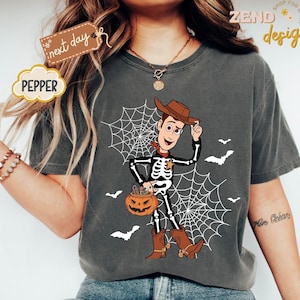 K&ouml;nnte beinhalten: Dunkelgraues T-Shirt mit Halloween-Design. Die Grafik zeigt einen Skelett-Cowboy mit einem K&uuml;rbis, vor einem Spinnennetz und Flederm&auml;usen. Das Wort "PEPPER" befindet sich in einem wolkenf&ouml;rmigen Design.