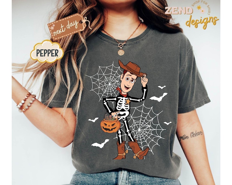 K&ouml;nnte beinhalten: Dunkelgraues T-Shirt mit Halloween-Design. Die Grafik zeigt einen Skelett-Cowboy mit einem K&uuml;rbis, vor einem Spinnennetz und Flederm&auml;usen. Das Wort "PEPPER" befindet sich in einem wolkenf&ouml;rmigen Design.