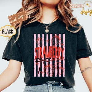 May include: Black t-shirt with a strawberry design. The design features the word "STRAWBERRY" in red, with pink vertical stripes and strawberry illustrations. The shirt also has the text "strawberry festival" and "good time".