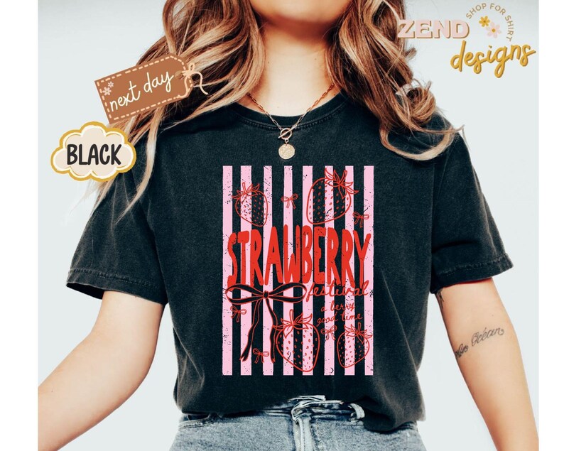 May include: Black t-shirt with a strawberry design. The design features the word "STRAWBERRY" in red, with pink vertical stripes and strawberry illustrations. The shirt also has the text "strawberry festival" and "good time".
