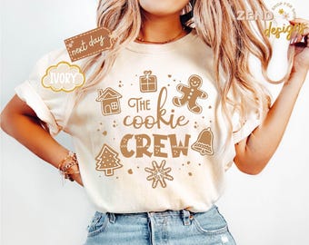 May include: Ivory-colored t-shirt with the text "The Cookie Crew" and gingerbread-themed graphics. The design includes a gingerbread man, house, gift, Christmas tree, bell, and snowflake. The shirt is worn by a person in blue jeans.