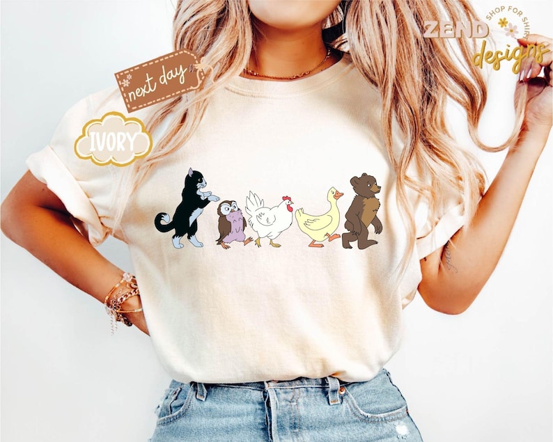 May include: Ivory-colored t-shirt featuring a whimsical cartoon design of a black and white cat, an owl, a white chicken, a yellow duck, and a brown bear walking in a line. The shirt also has the text "next day" and "Ivory".