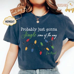 Pode incluir: Camiseta azul marinho com a frase "Probably just gonna Jingle some of the way. I'm tired." e uma sequ&ecirc;ncia de luzes de Natal coloridas. A camiseta tamb&eacute;m tem as palavras "next day" e "MIDNIGHT" em uma etiqueta.