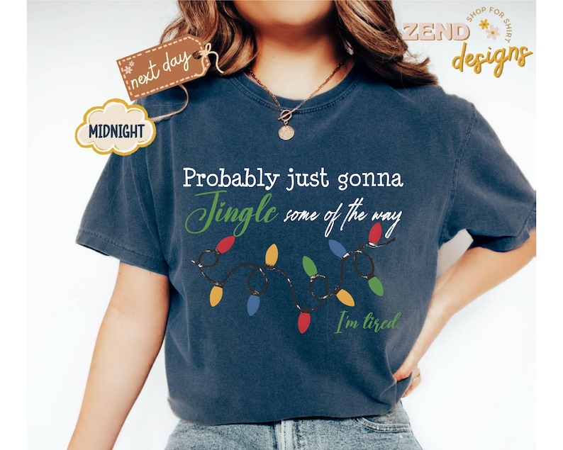 Pode incluir: Camiseta azul marinho com a frase "Probably just gonna Jingle some of the way. I'm tired." e uma sequ&ecirc;ncia de luzes de Natal coloridas. A camiseta tamb&eacute;m tem as palavras "next day" e "MIDNIGHT" em uma etiqueta.